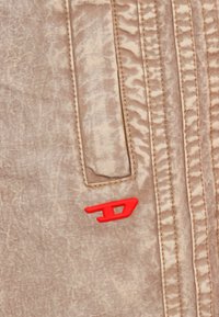 Μπεζ υφασμάτινη υφή με διακριτική γυαλάδα, που διαθέτει ραμμένες γραμμές και μια μικρή κόκκινη λεπτομέρεια λογότυπου. Μία τσέπη ορατή στην αριστερή πλευρά.