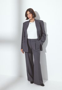 Harmaa räätälöity puku, jossa on kaksirivinen takki ja leveät housut. Valkoinen paita alla, sileä materiaali, minimaalinen varustus ja siistit linjat.