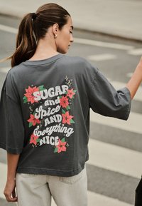 Szürke túlméretezett póló hátulján piros virágokat és a "SUGAR and SPICE AND Everything NICE" feliratot tartalmazó, vastag, fehér betűkkel írt grafika látható.