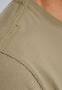 Camisa de algodón verde oliva que muestra una textura suave, con un cuello redondo y costuras de contraste sutiles a lo largo de la costura del hombro.
