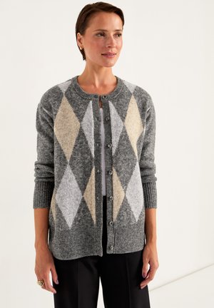 Vrouw in een grijze gebuttonte cardigan met een beige en witte ruitjespatroon, gecombineerd met zwarte broek, staand tegen een effen witte achtergrond.