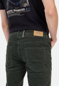 Dunkelgrüne Cordhosen mit einer strukturierten Oberfläche, ausgestattet mit einem Leder-Brandpatch und Gesäßtaschen, kombiniert mit einem schwarzen T-Shirt mit Grafikdesign.