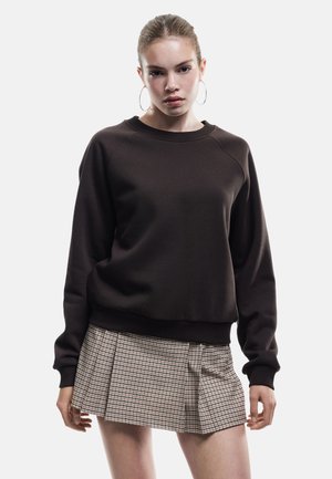 Brun sweatshirt med ribbat rundhals och långa ärmar, tillsammans med en veckad beige och brun rutig kjol. Slät tygtextur.