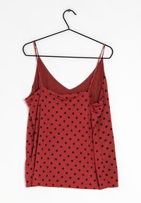 Haut sans manches rouge avec des pois noirs, fines bretelles réglables, accroché à un cintre noir sur fond blanc.