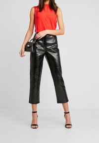 Haut rouge sans manches avec un design à découpes en forme de goutte, assorti à un pantalon noir taille haute en simili-cuir effet crocodile. Le mannequin tient un petit sac à main noir.