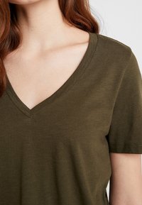 Olivově zelené tričko s výstřihem do V vyrobené z měkkého materiálu, s krátkými rukávy a volným střihem, hladkou texturou a jednoduchým designem.