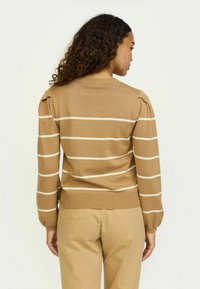 Maglione lavorato a maglia color marrone chiaro con strisce bianche, dotato di dettagli sulle spalle a sbuffo e fondo a coste. Modello che indossa pantaloni beige.