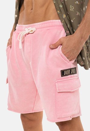 Homme portant un short cargo rose clair avec une taille à cordon et un patch noir "Von Dutch", les mains dans les poches, et une chemise à motifs partiellement ouverte.