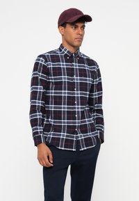 Camicia a quadri blu navy e bianca con accenti rossi, maniche lunghe e orlo arrotondato, abbinata a un cappellino bordeaux.