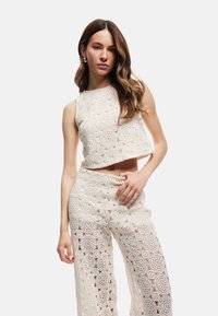 Conjunto de top y pantalones de crochet marfil con patrones circulares, diseño recortado y ajuste de pierna amplia. Textura transparente, adornos de costura decorativa en toda la prenda.