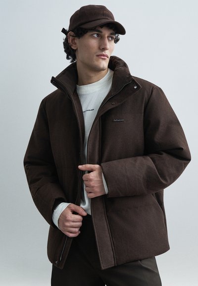 Braune gesteppte Jacke mit hohem Kragen und Reißverschluss, mit einem dezenten Logo auf der Brust. Getragen über einem weißen Rundhalsshirt.