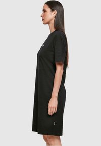 Czarna bawełniana sukienka t-shirt z krótkim rękawem, luźny krój, długość do kolan oraz małe logo z przodu, przedstawiająca prosty design.