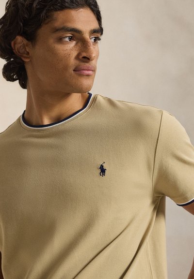 Μπεζ κοντομάνικο πόλο με εφαρμοστή γραμμή, στρογγυλή γιακά με περιθώριο σε ναυτικό και ανοιχτό μπλε, και κεντημένο λογότυπο σε ναυτικό χρώμα στο στήθος.