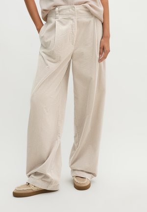 Beige wijde corduroy broek gedragen met beige instapschoenen met bontafwerking, getoond op een staand persoon tegen een effen achtergrond.