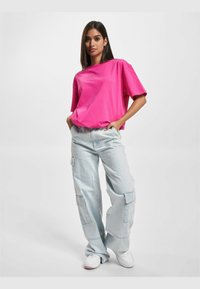Camiseta de gran tamaño de color rosa brillante con un logo, combinada con pantalones cargo de color azul claro que cuentan con bolsillos con parche. El modelo lleva zapatillas blancas.