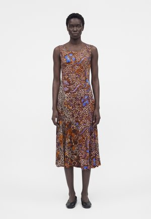 Kvinde stående iført en ærmeløs, mellemlang kjole med brune, blå og hvide abstrakte blomster- og prikmønstre, kombineret med sorte sko.