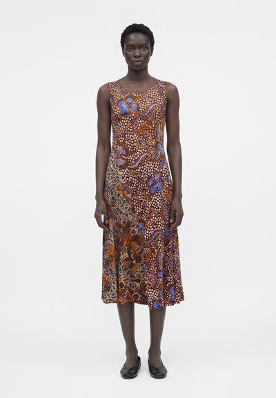 Femme debout portant une robe sans manches, mi-longue, avec des motifs floraux abstraits et à pois marron, bleu et blanc, associée à des chaussures noires.