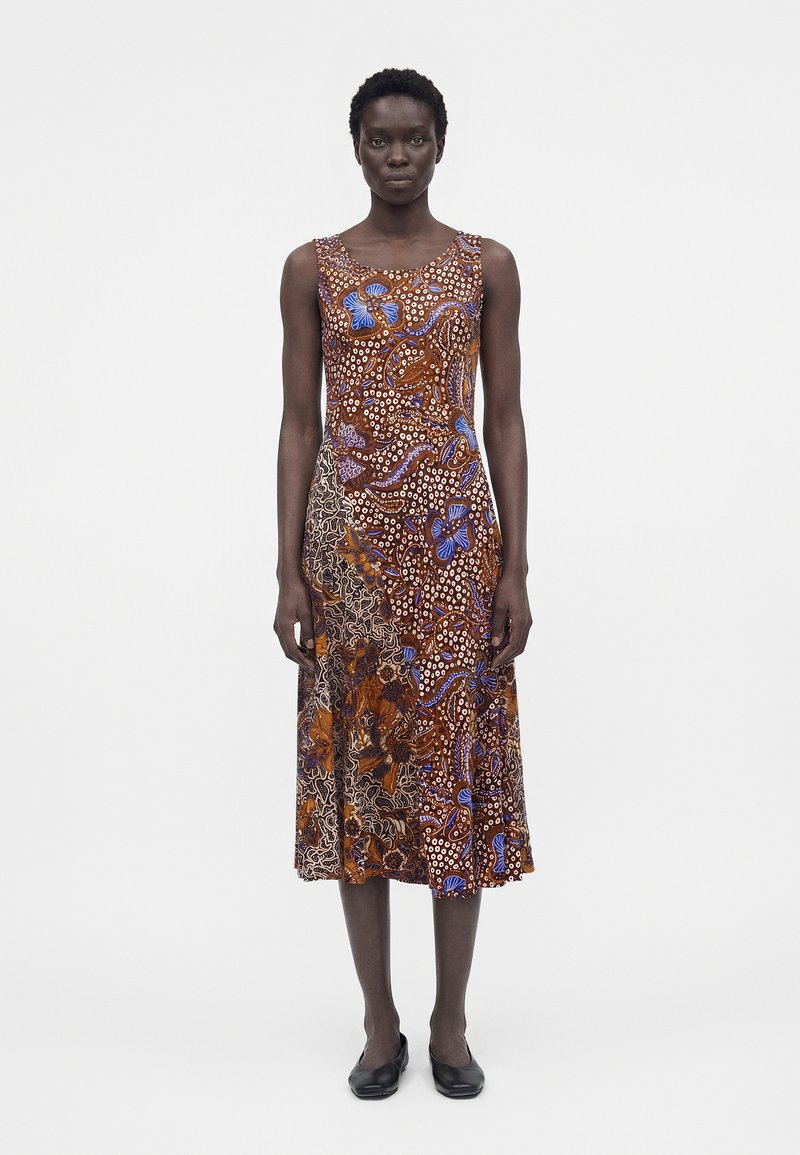 Γυναίκα που στέκεται, φορώντας αμάνικο, μεσαίου μήκους φόρεμα με καφέ, μπλε και λευκά αφηρημένα λουλουδένια και πουα μοτίβα, συνδυασμένο με μαύρα παπούτσια.