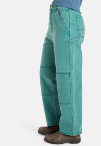 Pantalon en denim turquoise avec une coupe droite, présentant des coutures visibles et des poches latérales, porté avec des sabots marron.