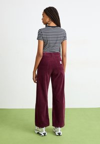 Pantaloni in velluto a coste bordeaux a vita alta con gambe larghe, abbinati a una maglietta aderente a righe bianche e nere. Modella di spalle, con sneakers argentate.