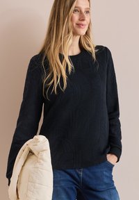 Marineblauer strukturierter Pullover mit einem subtilen Blättermuster, Rundhalsausschnitt und langen Ärmeln, kombiniert mit einer hellen gesteppte Tasche.