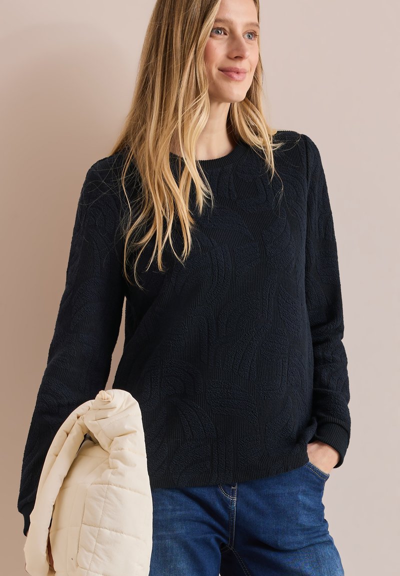 Marineblauer strukturierter Pullover mit einem subtilen Blättermuster, Rundhalsausschnitt und langen Ärmeln, kombiniert mit einer hellen gesteppte Tasche.