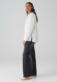 Camicia bianca a maniche lunghe con una vestibilità comoda e spacchi laterali, abbinata a jeans dritti scuri e scarpe bianche con strisce rosse.