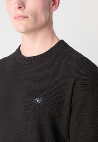 Melns teksturēts adīts džemperis ar ribotu apkakli un mazu logotipa etiķeti uz krūtīm. Audums izskatās mīksts un biezs.