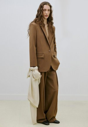 Osoba oblečená v příliš velkém hnědém obleku drží béžový šátek, stojící před jednoduchým bílým pozadím stěny a podlahy.