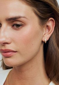 Guld hoops øreringe med en tekstureret overflade, båret i højre øre, der komplementerer glat hud og neutral makeup. Enkel kædehalssmykke synlig.