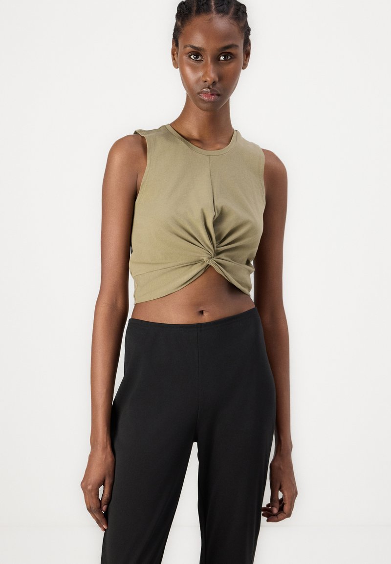 Olivgrünes, ärmelloses Cropped-Top mit einer verdrehten Vorderseite, kombiniert mit schwarzen, hochgeschnittenen Hosen. Glatte Textur insgesamt.