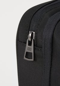 Black fabric bag corner with textured surface, featuring a black zipper and metallic pull tab, showing intricate stitching detail.