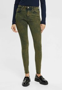 Jeans skinny de color verde oliva con una textura desgastada, diseño de cinco bolsillos y cierre de botón. Combinados con mocasines negros y brillantes.
