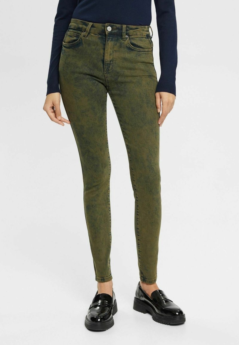 Jeans skinny de color verde oliva con una textura desgastada, diseño de cinco bolsillos y cierre de botón. Combinados con mocasines negros y brillantes.