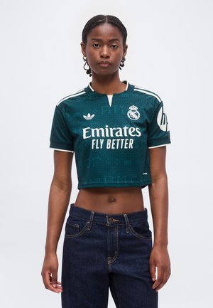Jeune femme portant un maillot de football Real Madrid court vert foncé avec le logo blanc « Emirates Fly Better », assorti à un jean bleu foncé.