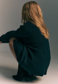 Ciemnozielony, oversize'owy sweter z dzianiny z detalem guzików, zestawiony z czarnymi botkami z frędzlami sięgającymi kostki. Modelka siedzi, prezentując układ i teksturę ubrania.