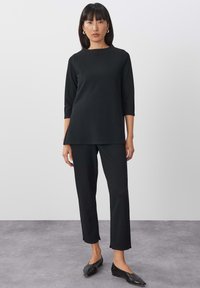 Zwarte top met driekwartmouwen en bijpassende broek, gemaakt van een gladde stof. De slim-fit broek heeft een iets verkorte lengte. Platte schoenen completeren de outfit.