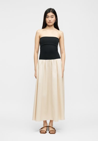 Νεαρή γυναίκα με μακριά σκοτεινά μαλλιά, που φοράει φορέματα με μαύρο κορσέ χωρίς τιράντες και μπεζ φούστα, με καφέ σανδάλια, στέκεται μπροστά σε απλό φόντο.