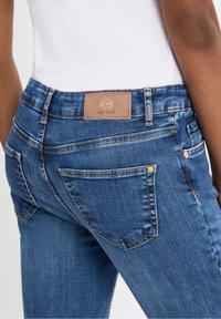 Vaqueros de mezclilla en azul oscuro con una textura suave. Presenta una etiqueta de piel en la cintura y cinco bolsillos, incluyendo dos bolsillos traseros.