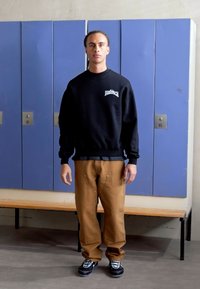 Black sweatshirt with logo, beige pants, and black sneakers. Background features blue lockers and a wooden bench. Simple, casual outfit.
