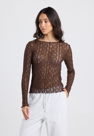 Vrouw in een bruin kantenshirt met lange mouwen en lichtgrijze joggingbroek, staand met één hand in de zak tegen een effen achtergrond.
