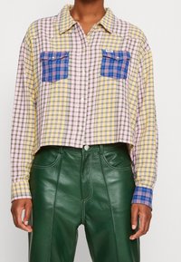 Gekürztes Karoshemd mit gelben, rosa und grünen Karos; verfügt über zwei blaue Karotaschen und einen Knopfverschluss. Kombiniert mit grünen Lederhosen.