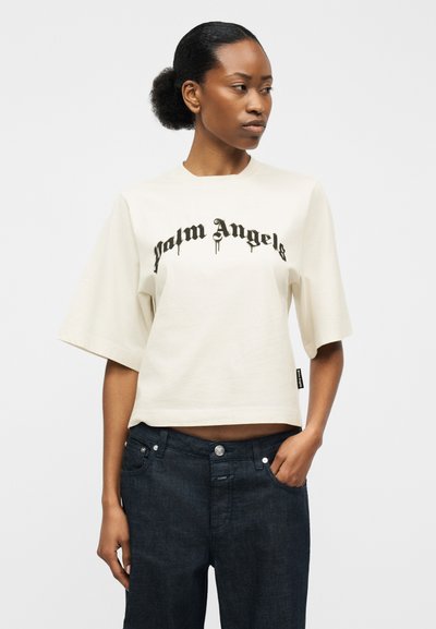 Žena s vlasmi v drdole, oblečená v bielom krátkom tričku s nápisom "palm Angels" a tmavých džínsoch, stojaca s jednou rukou v vrecku.