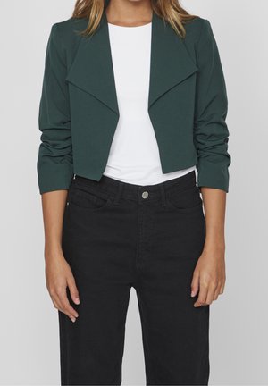 Femme portant une veste courte vert foncé aux manches froncées sur un haut blanc et un jean noir taille haute, devant un fond uni.