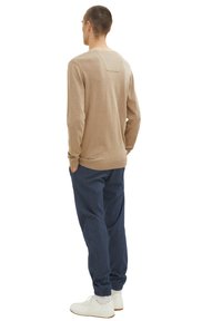 Beiger Strickpullover mit langen Ärmeln und einem quadratischen Rückendetail, kombiniert mit dunkelblauen lockeren Hosen und weißen Turnschuhen.