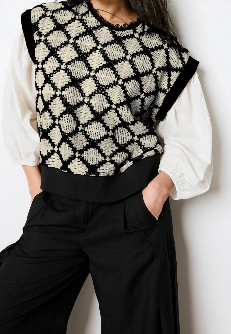 Personne portant un pull sans manches à motif noir et blanc sur une blouse blanche à manches longues bouffantes avec un pantalon noir, mains dans les poches.