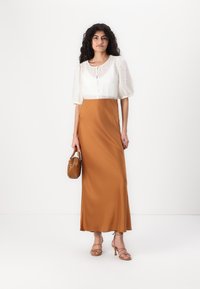 Abito maxi in satin marrone con una blusa bianca texturizzata caratterizzata da maniche a sbuffo e chiusura con bottoni; il modello tiene una piccola borsa marrone.