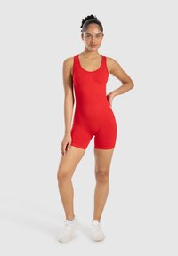 Mono rojo sin mangas fabricado en tejido elástico, con un diseño ajustado que termina a mitad del muslo. La modelo lleva zapatillas deportivas blancas.