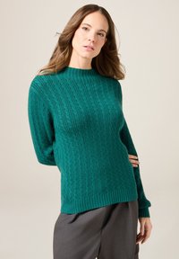 Türkis gestrickter Pullover mit hohem Kragen, der über eine gerippte Textur und ein Zopfmuster verfügt. Lockere Ärmel sowie gerippte Bündchen und Saum.
