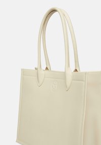 Beżowa torba na ramię z podwójnymi uchwytami, gładką fakturą, wytłoczonym logo na przodzie oraz przeszytą obwódką, charakteryzująca się sztywnym kształtem.